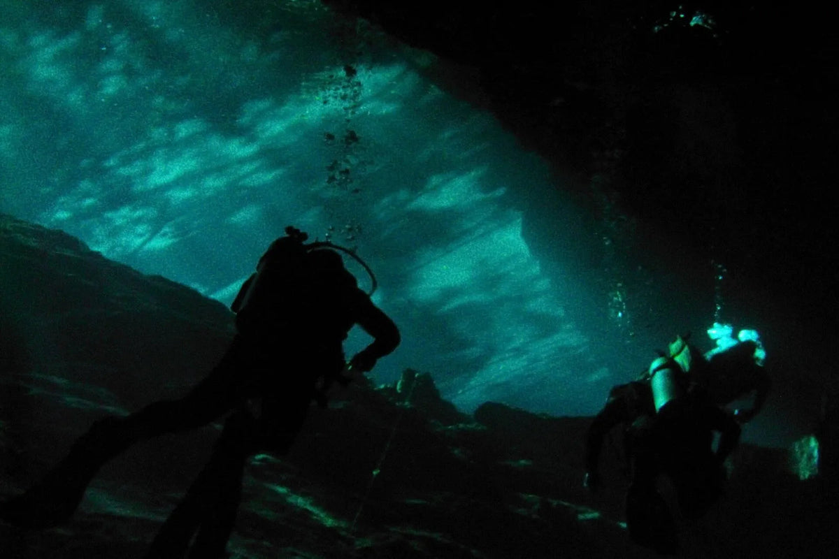 Zwei Taucher als Schattenriss unter einem Felsdach, durch die Öffnung darüber fällt grünes Licht ins Wasser