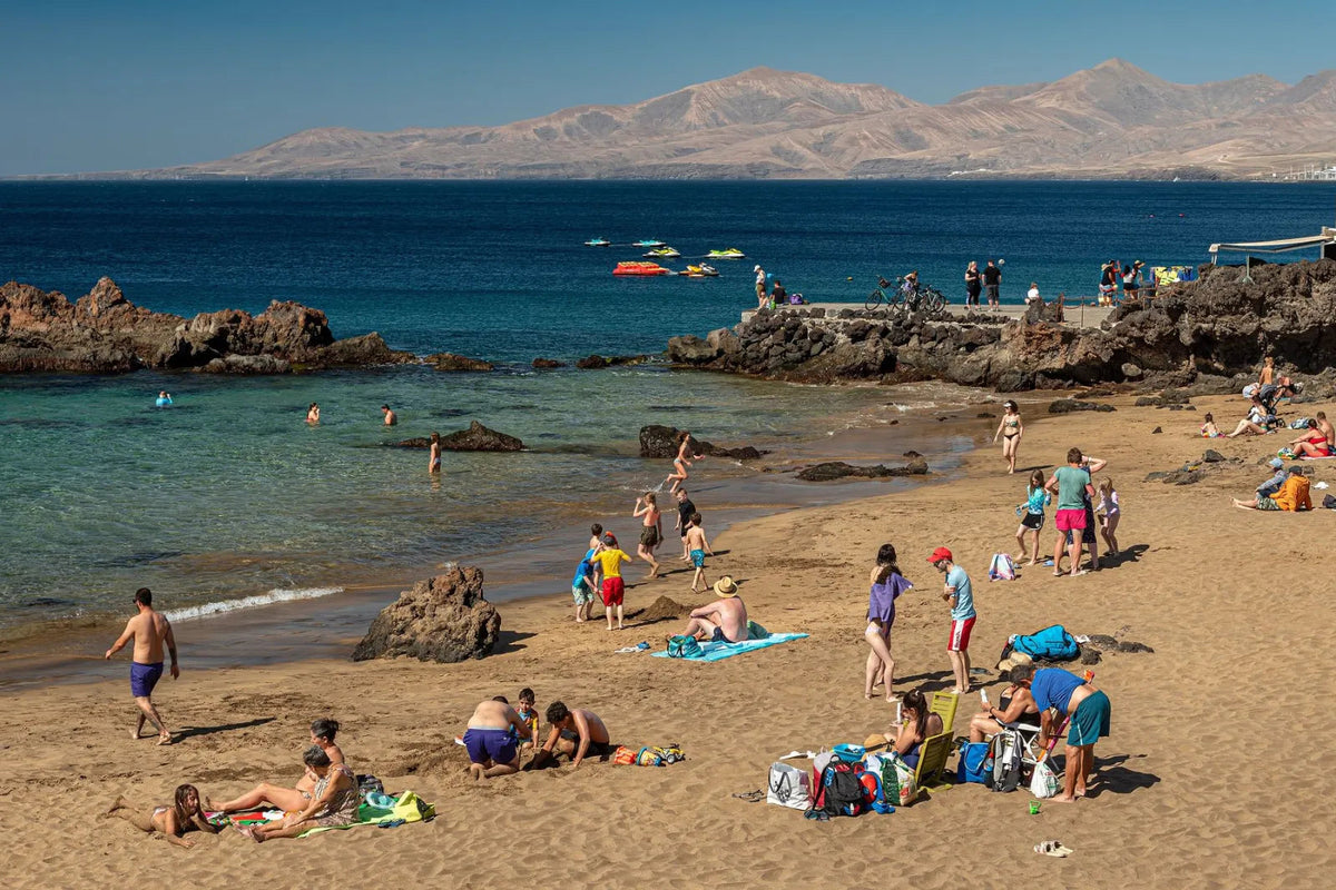 Sandbucht der Playa Chica mit Badegästen, links und rechts schwarze Lavafelsen im klaren Wasser, am Horizont Berge