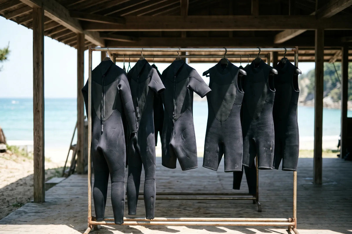 Lange und kurze schwarze Neoprenanzüge hängen an einer Stange unter einem Holzdach, dahinter Strand und Meer