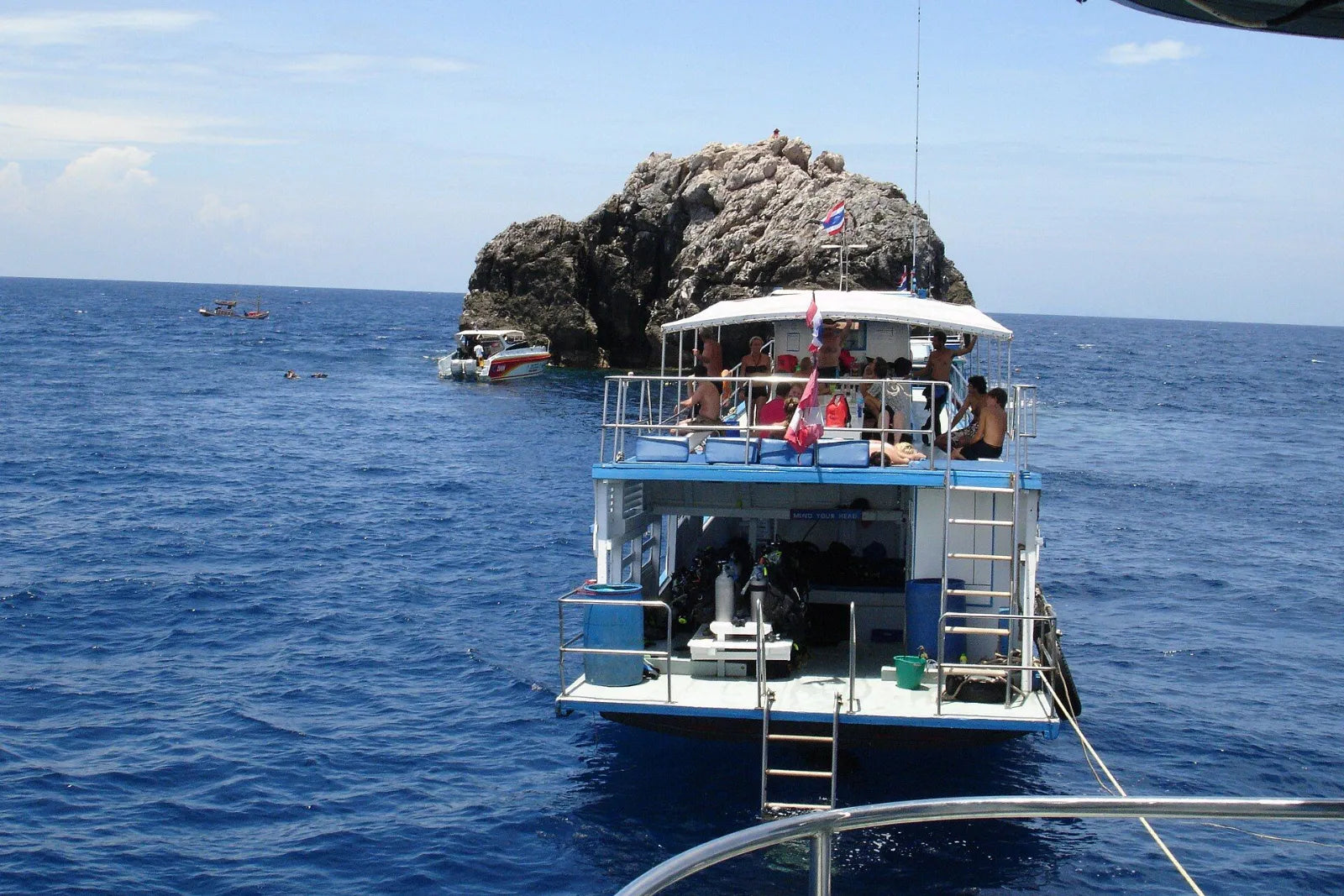 Dive boat with guests on deck in front of a gray rock, which rises steeply from the deep blue sea