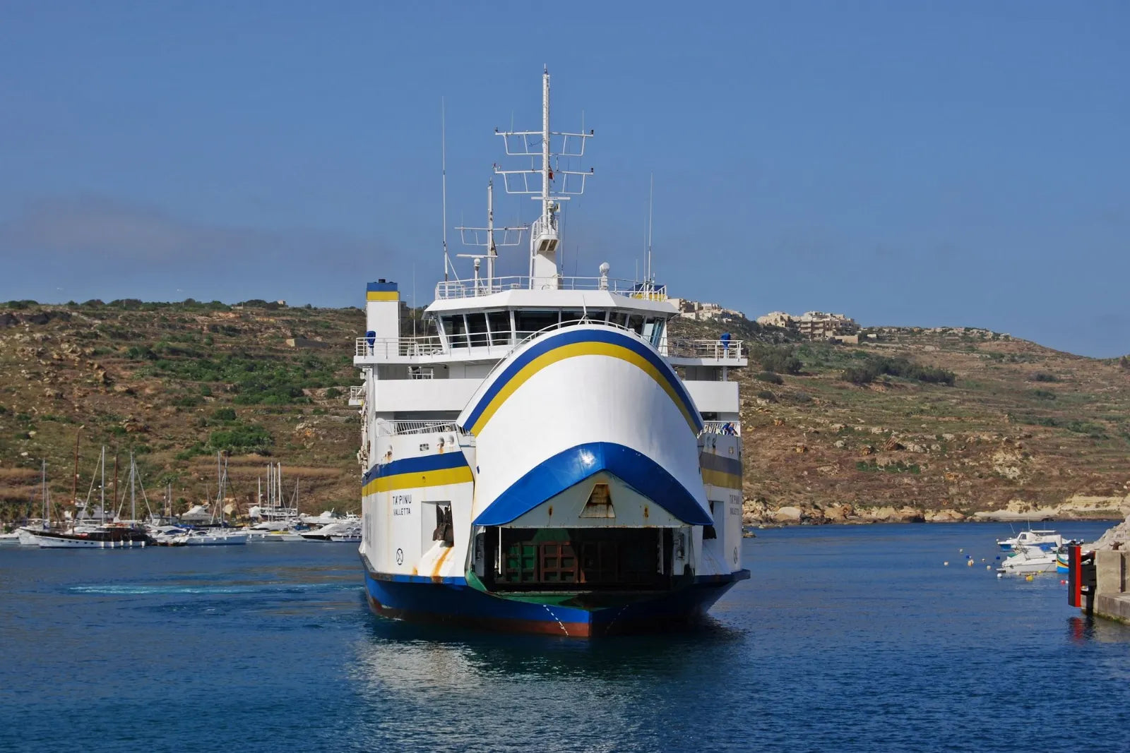 The Ta' Pinu car ferry with its raised bow visor departs from the port of Mġarr on Gozo, with green hills and a marina behind it