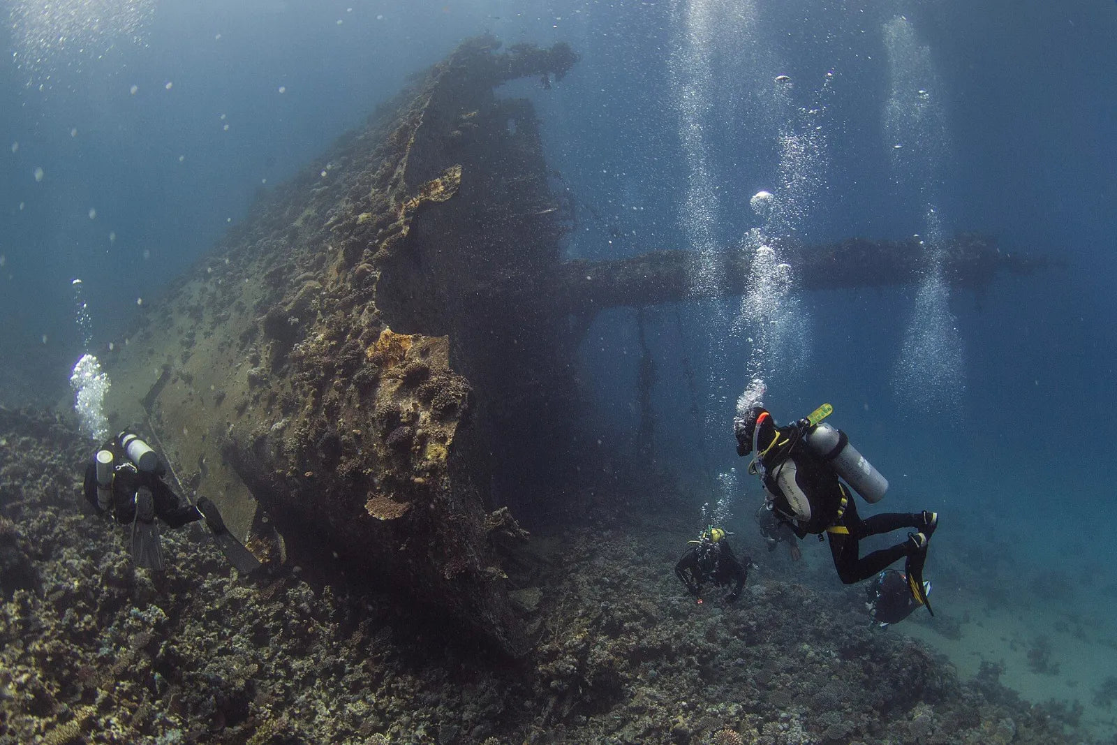 Two divers at the overgrown bow of a wreck on a rocky bottom, with a mast and cargo boom behind them
